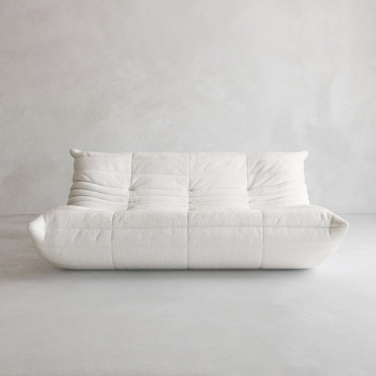 The Rolo Lounge Three Seater, a modern white armless sofa with plush tufting and mid-century inspiration, sits against a light gray wall and floor, creating a minimalist and cozy atmosphere.