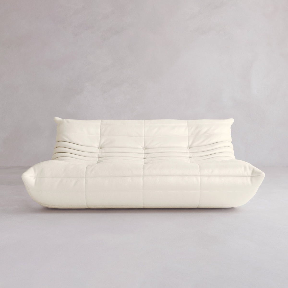 The Rolo Lounge Three Seater, a modern white sofa with a plush tufted design and gentle horizontal folds, sits against a light gray wall and floor for a cozy, minimalist space inspired by mid-century style.