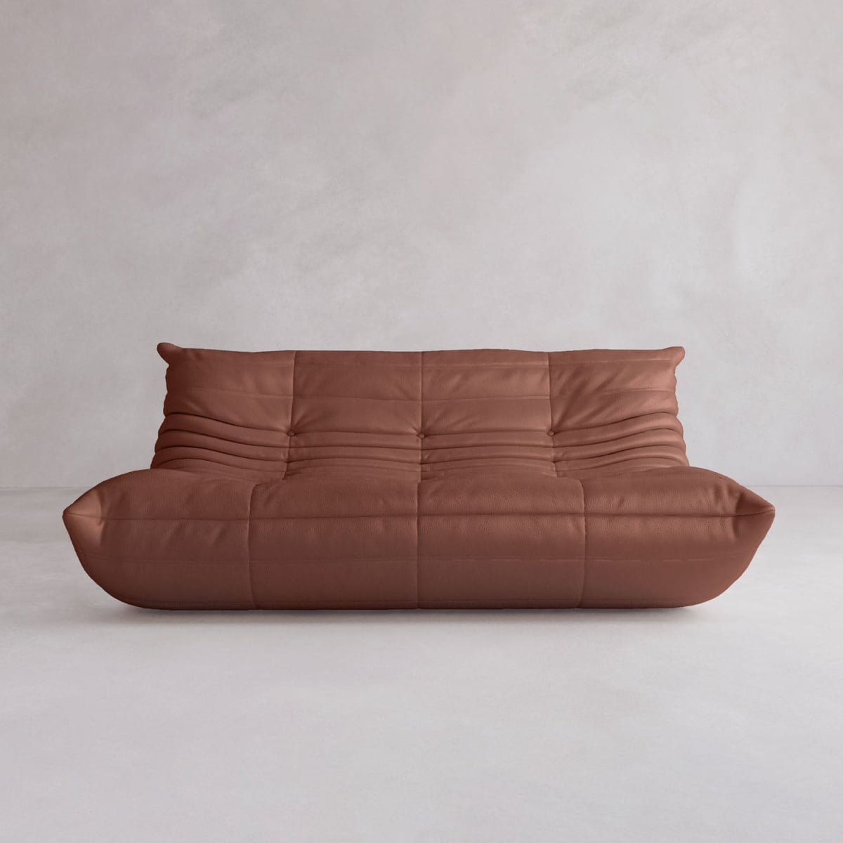 The Rolo Lounge Three Seater is a modern brown leather sofa with a low profile, deep seat, and soft quilted upholstery. It features subtle mid-century design elements and sits against a light-colored wall and floor.