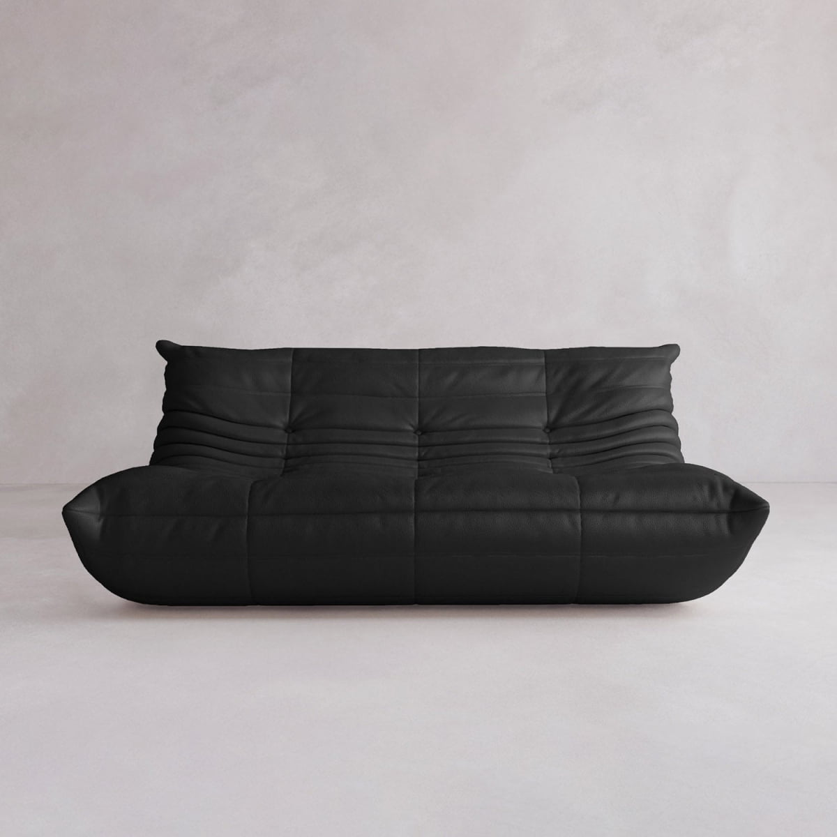 The Rolo Lounge Three Seater, a modern black leather sofa with soft tufted cushions and a low profile inspired by mid-century style, sits against a light gray textured wall on a pale floor.