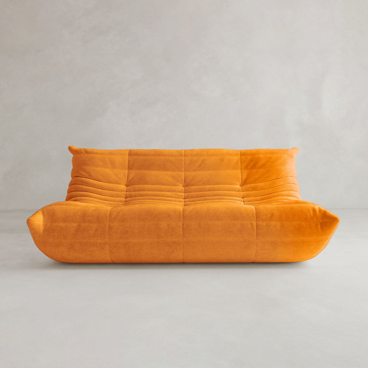The Rolo Lounge Three Seater, a modern armless orange sofa with plush quilting and mid-century inspiration, sits on a plain light gray floor against a light gray wall.