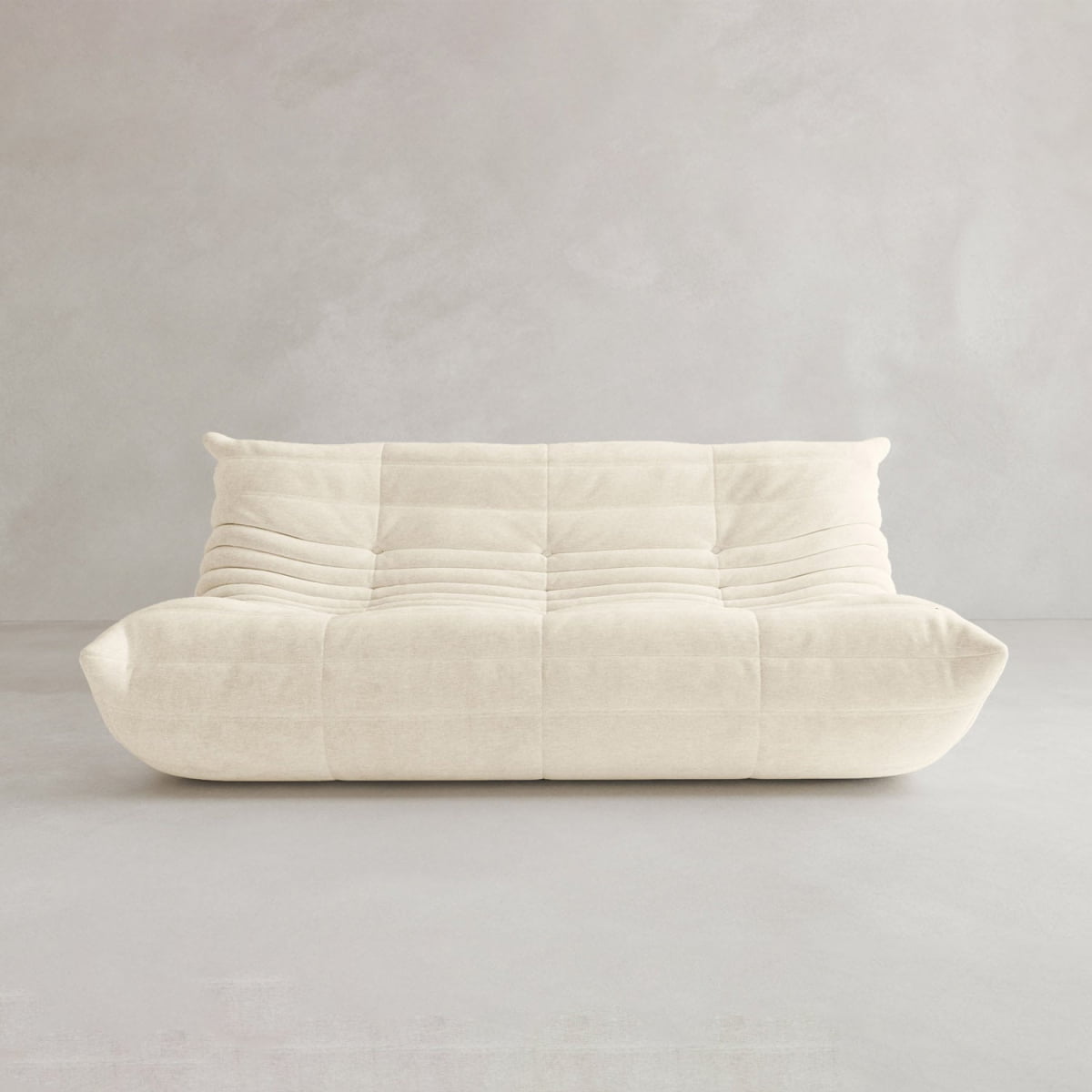 The Rolo Lounge Three Seater is a modern cream-colored, low-profile sofa featuring a plush tufted design and soft, rounded edges inspired by mid-century style, displayed against a plain light gray background.