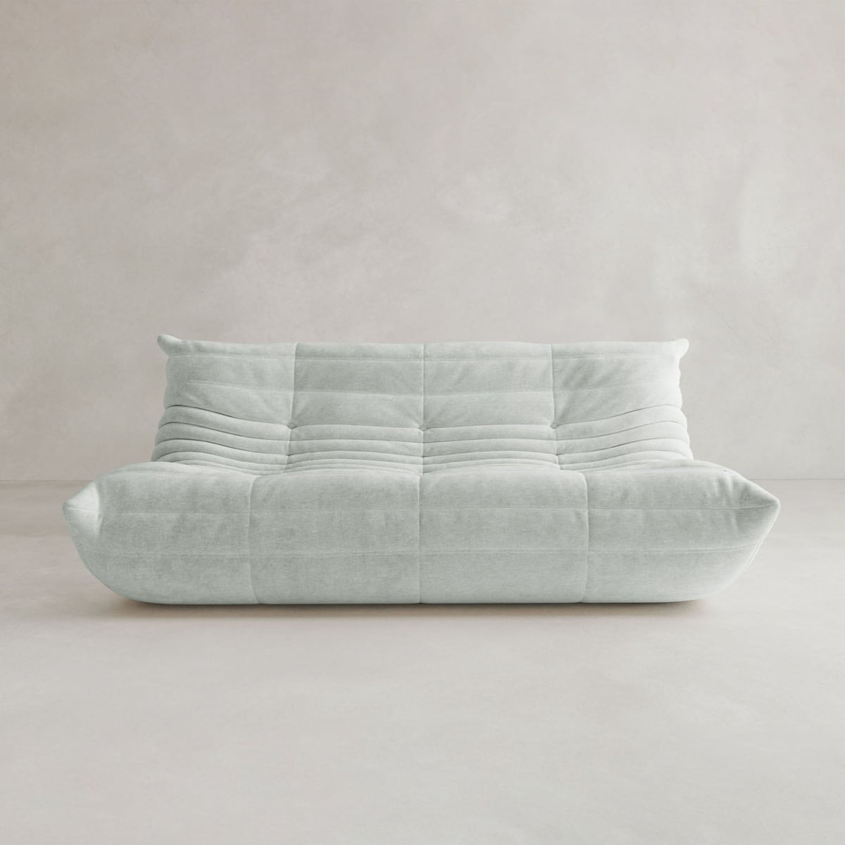 The Rolo Lounge Three Seater is a modern, light gray armless sofa with tufted, quilted details and a low backrest inspired by mid-century design, set against a plain, light-colored wall and floor.