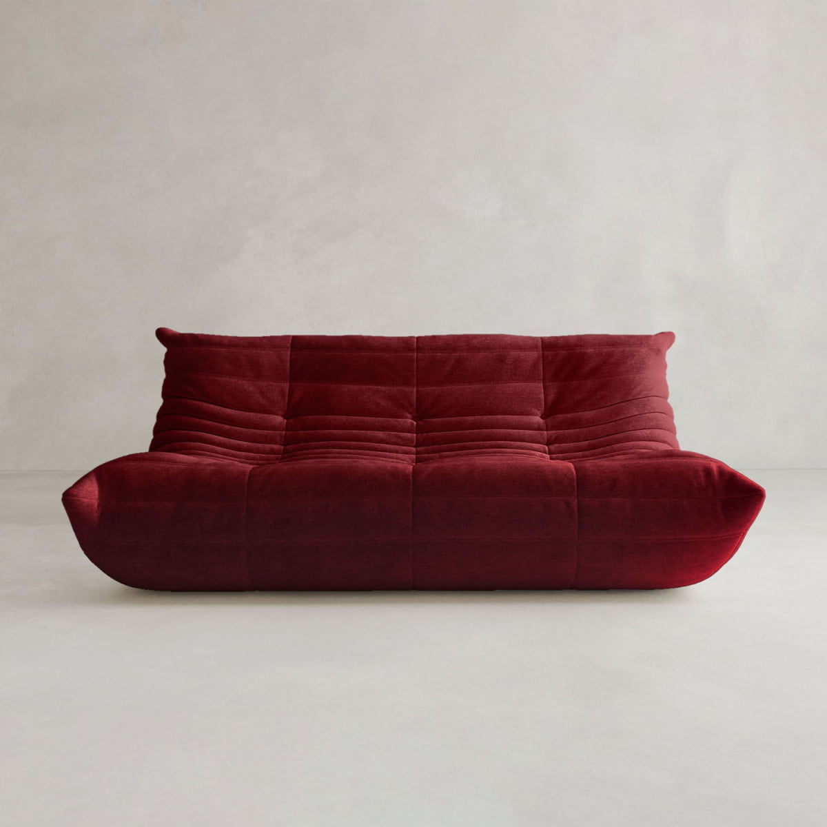 The Rolo Lounge Three Seater, a deep red velvet sofa with tufted cushions and a minimalist mid-century design, features a low profile and is set against a light gray textured background.