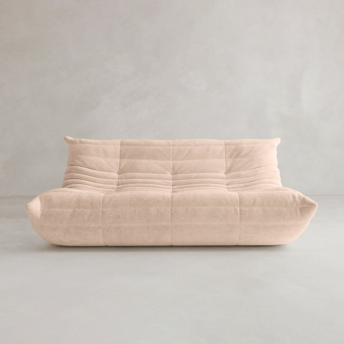 The Rolo Lounge Three Seater is a beige contemporary modular sofa with a low backrest, deep seat, and soft quilted upholstery, shown against a plain light gray wall and floor.