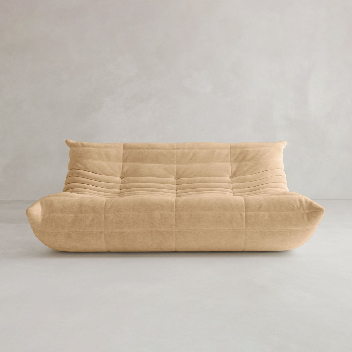 The Rolo Lounge Three Seater, a modern armless beige sofa with a quilted cushioned design inspired by mid-century style, sits against a plain light gray wall and floor.
