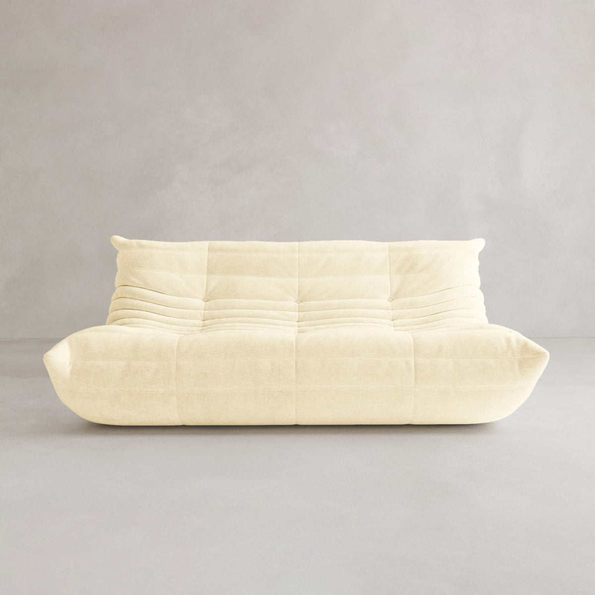 The Rolo Lounge Three Seater is a cream-colored, modern armless sofa with a soft quilted texture and low cushioned backrest, mid-century inspired, shown against a plain light gray background.