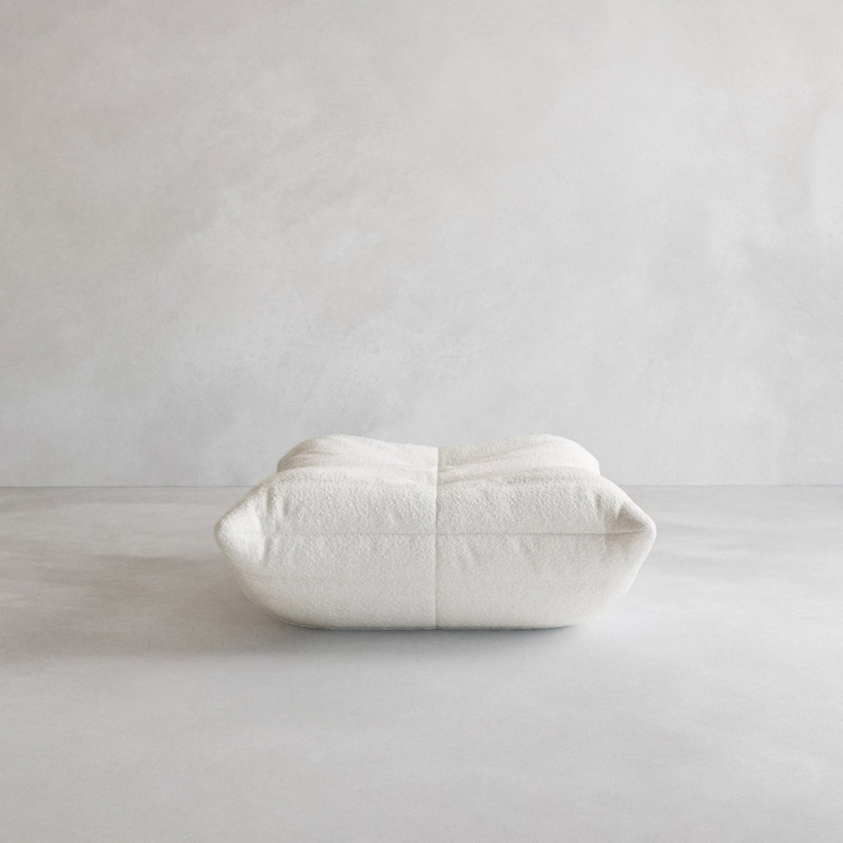The Rolo Ottoman, a large square white floor pillow with a cushioned top and soft textured surface, sits on a minimalist light gray floor against a plain wall—perfect for any modern space.