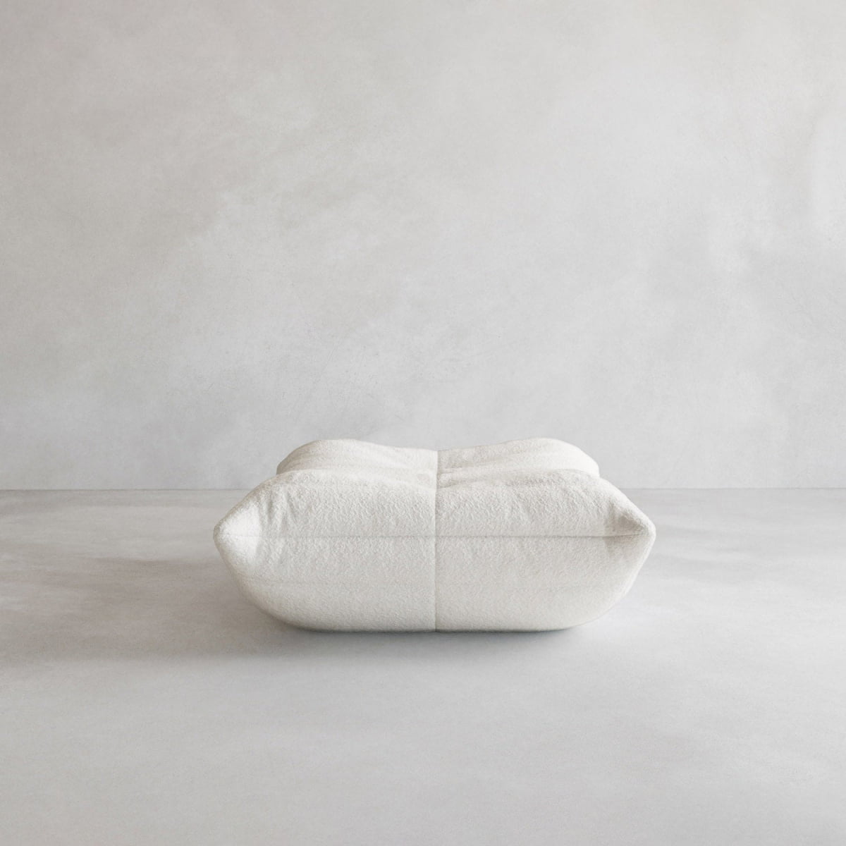 The Rolo Ottoman is a minimalist, square white floor cushion with a cushioned top, shown on a smooth light gray surface against a plain light gray wall—perfect for modern spaces.