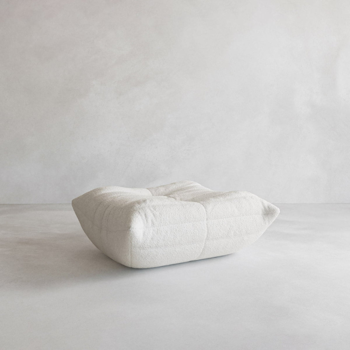 The Rolo Ottoman is a modern, white, square ottoman with a sleek design and cushioned top for plush comfort, shown on a light gray floor against a plain light-colored wall.
