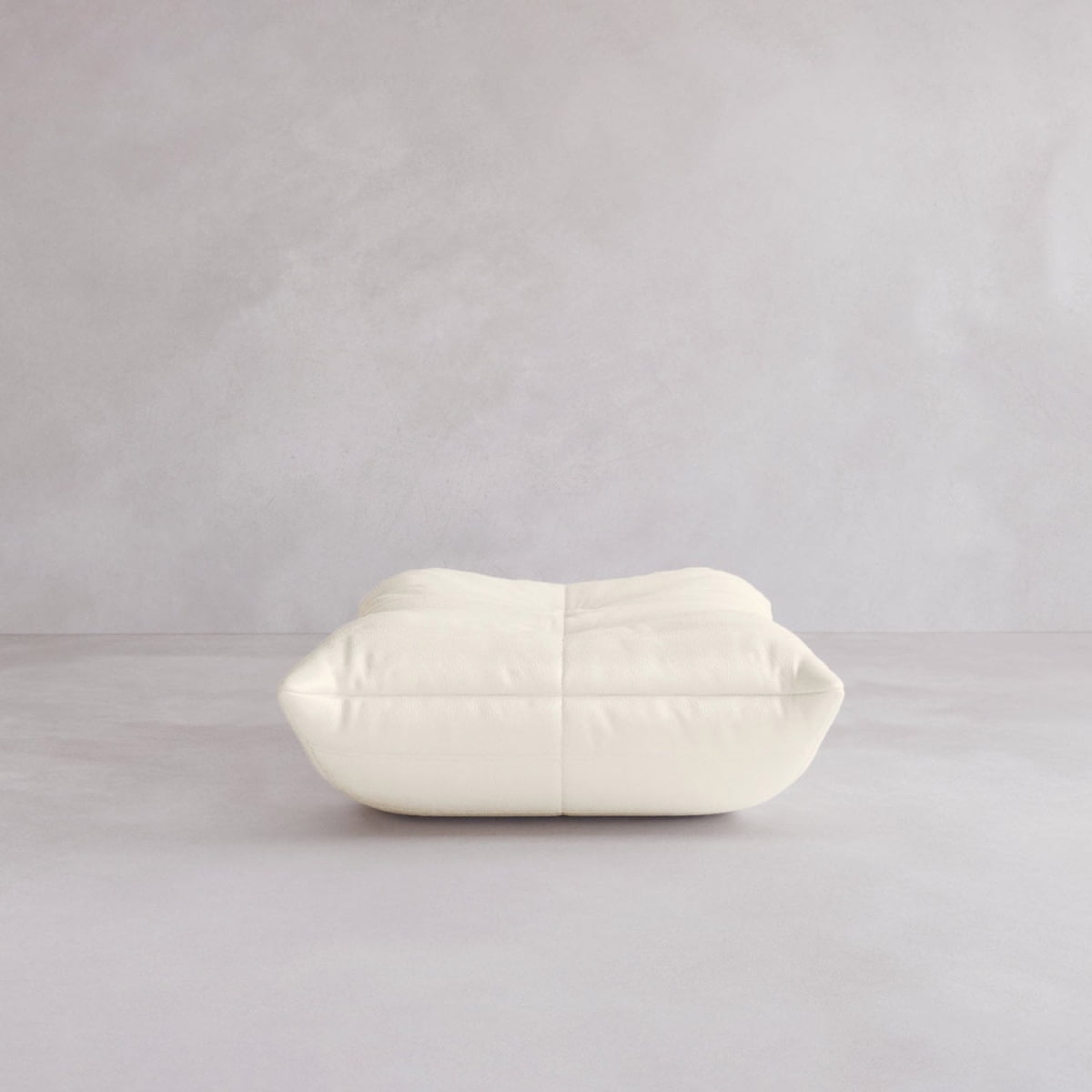 The Rolo Ottoman, a soft, rectangular, off-white cushioned piece, lies flat on a light neutral surface with a plain light gray wall in the background.