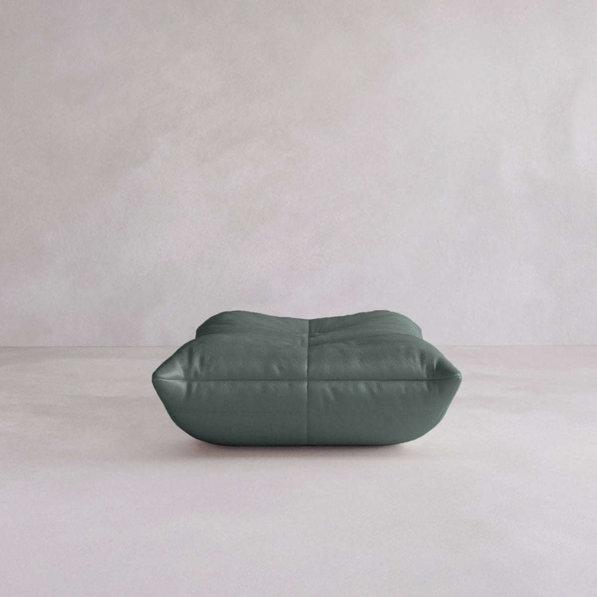 The Rolo Ottoman, a dark green square-shaped cushion with visible seams and a sleek design, rests on a light minimalist floor against a pale wall.