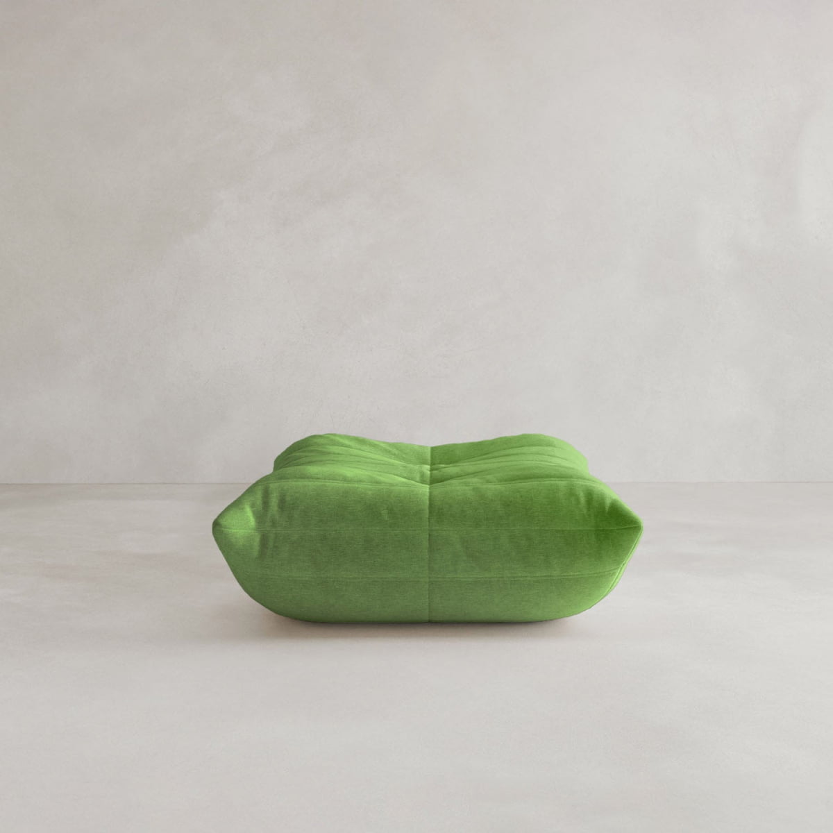 The green Rolo Ottoman, a plush, square floor cushion with visible seams and a cushioned top, sits on a light surface against a pale wall, showcasing its modern minimalist design.