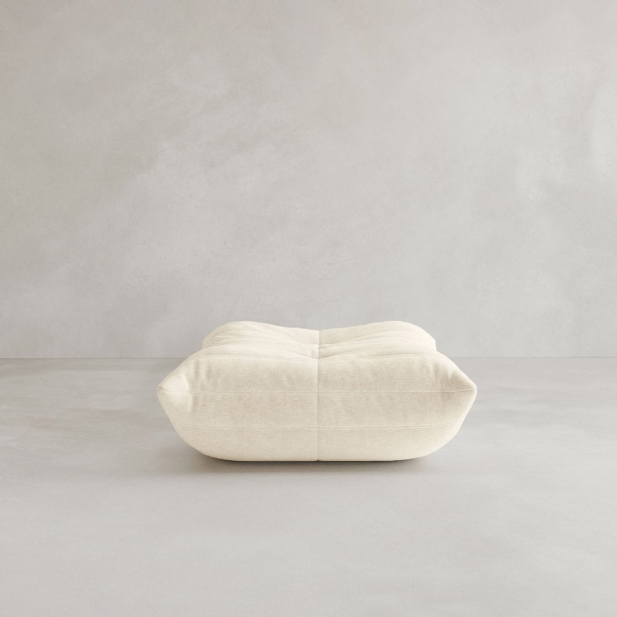 The Rolo Ottoman, a low, rectangular cream-colored floor cushion with soft, rounded edges and visible seams dividing its cushioned top, sits in an empty room with light gray textured walls and flooring.