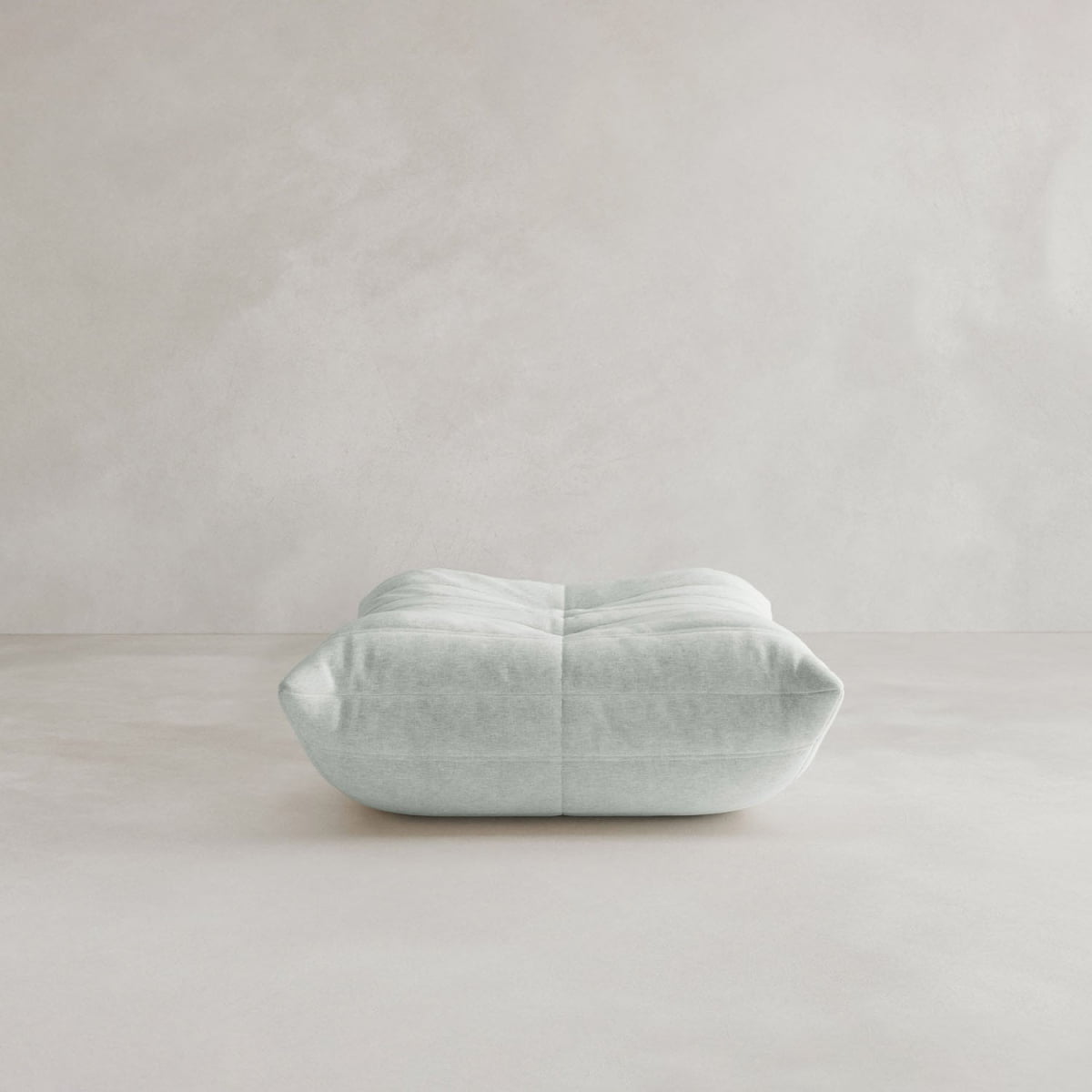 The Rolo Ottoman, a modern light gray floor cushion with a cushioned top and soft, tufted seams, sits on a smooth, light-colored floor against a plain, textured wall.