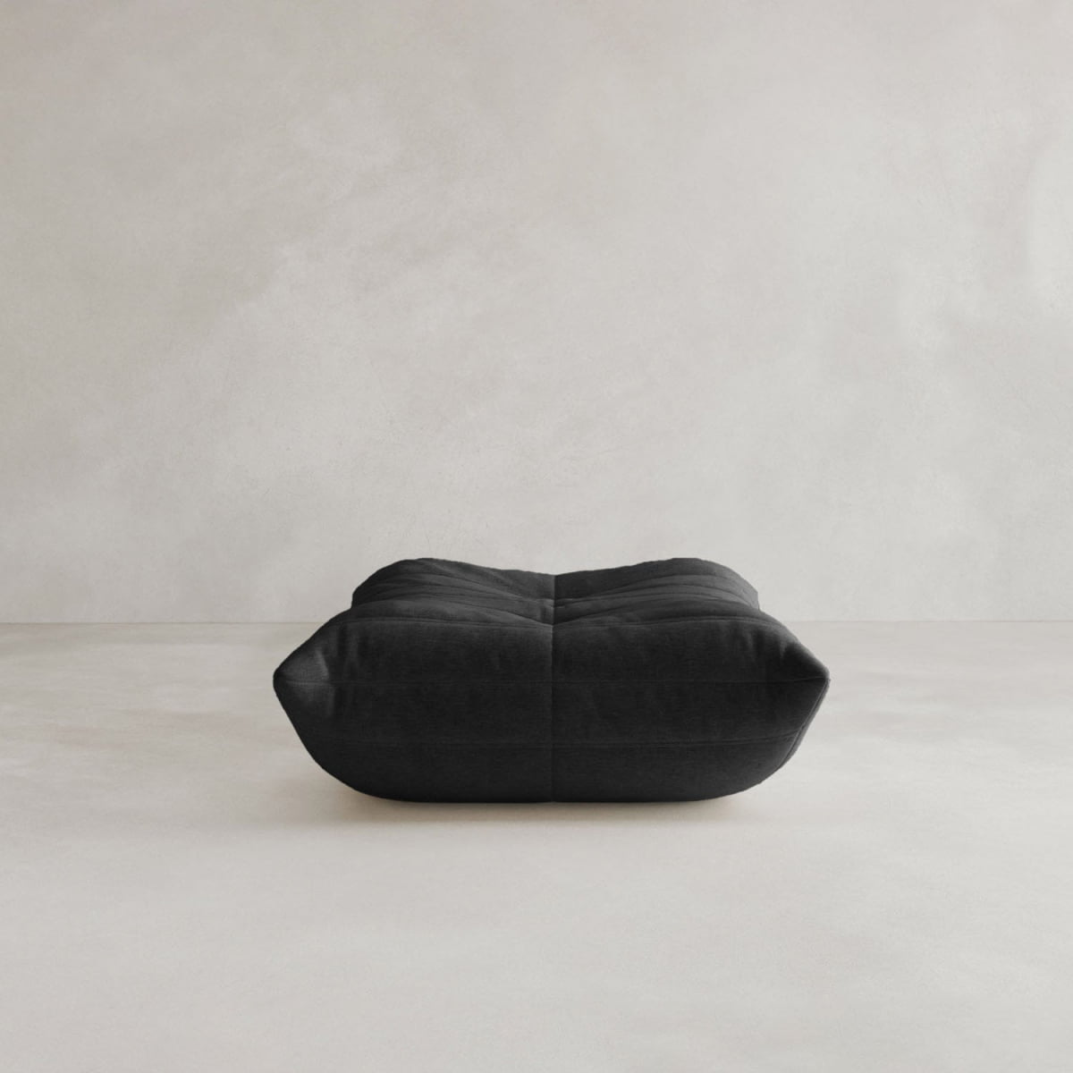 The Rolo Ottoman, a black, low-profile rectangular floor cushion with a cushioned top, rests on a light minimalist floor against a plain textured wall.
