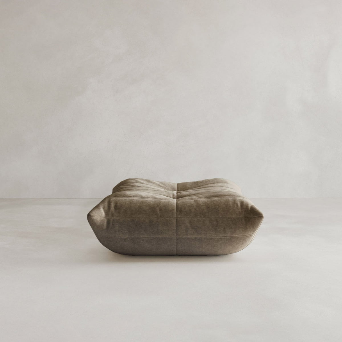 The Rolo Ottoman, a soft, square brown floor cushion with a cushioned top, sits alone on a neutral surface against an off-white background.