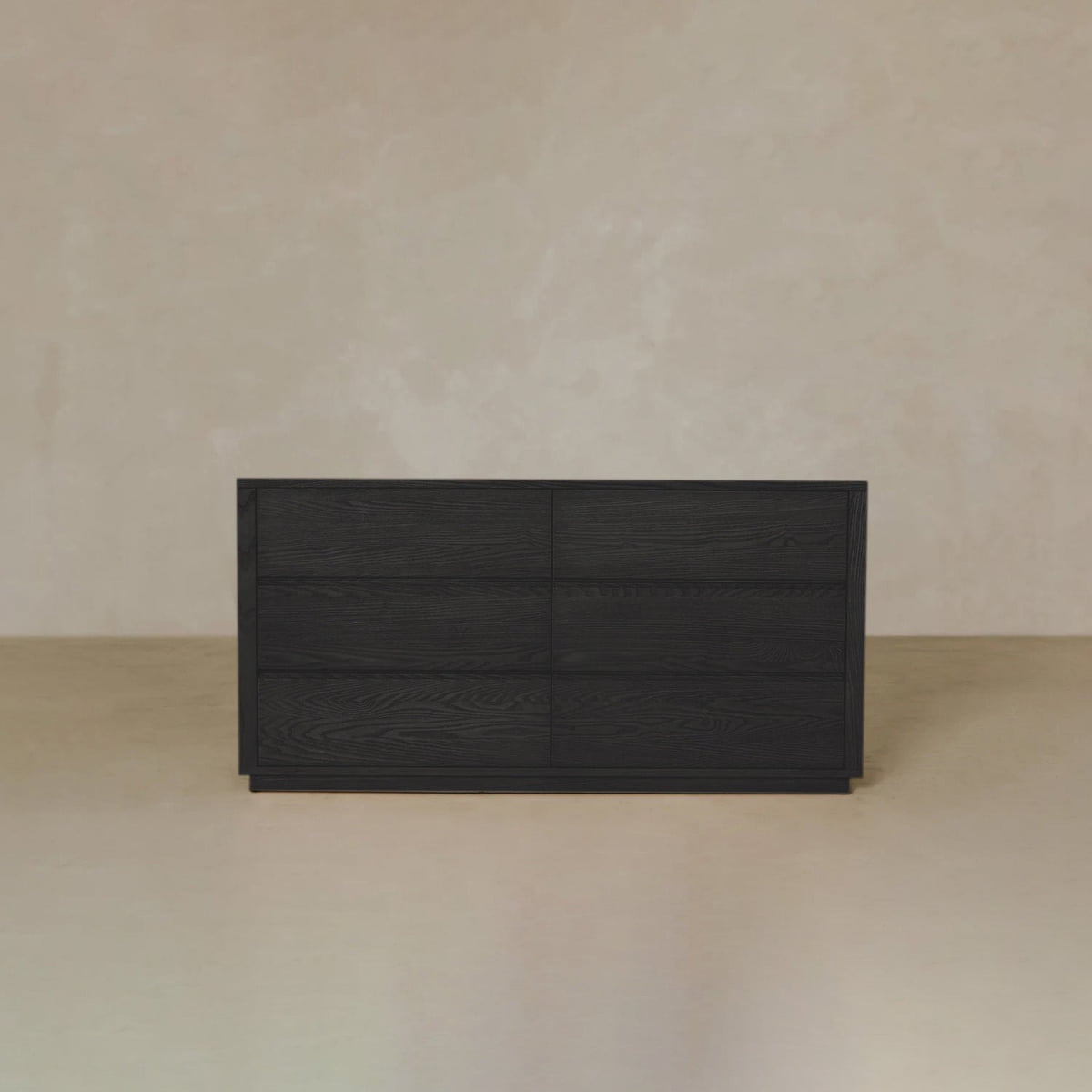 The Sandringham Dresser, a sleek black wood piece with six drawers, provides ample storage and style for minimalist bedrooms with plain beige walls and floors.