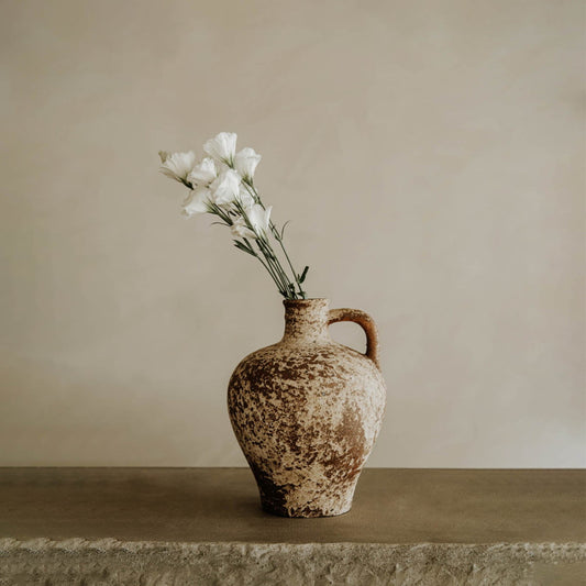The Sato vase, crafted from textured clay with a handle for an artisanal touch, holds white flowers against a neutral beige background—perfect as timeless home decor.