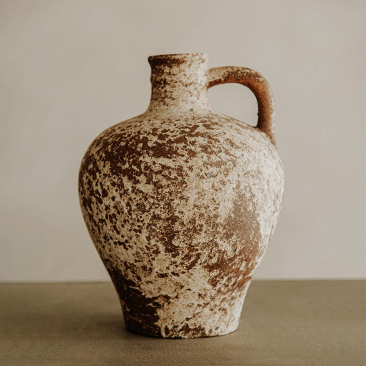 The Sato is a rustic clay jug with a single handle and slim neck, featuring a mottled brown and white finish—ideal for bringing artisan charm to your home decor against neutral backgrounds.