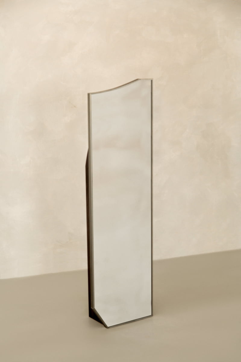 The Shard Mirror S, a tall and narrow mirror with a slightly irregular, angled top edge, brings modern elegance as it stands freestanding against a neutral beige background.