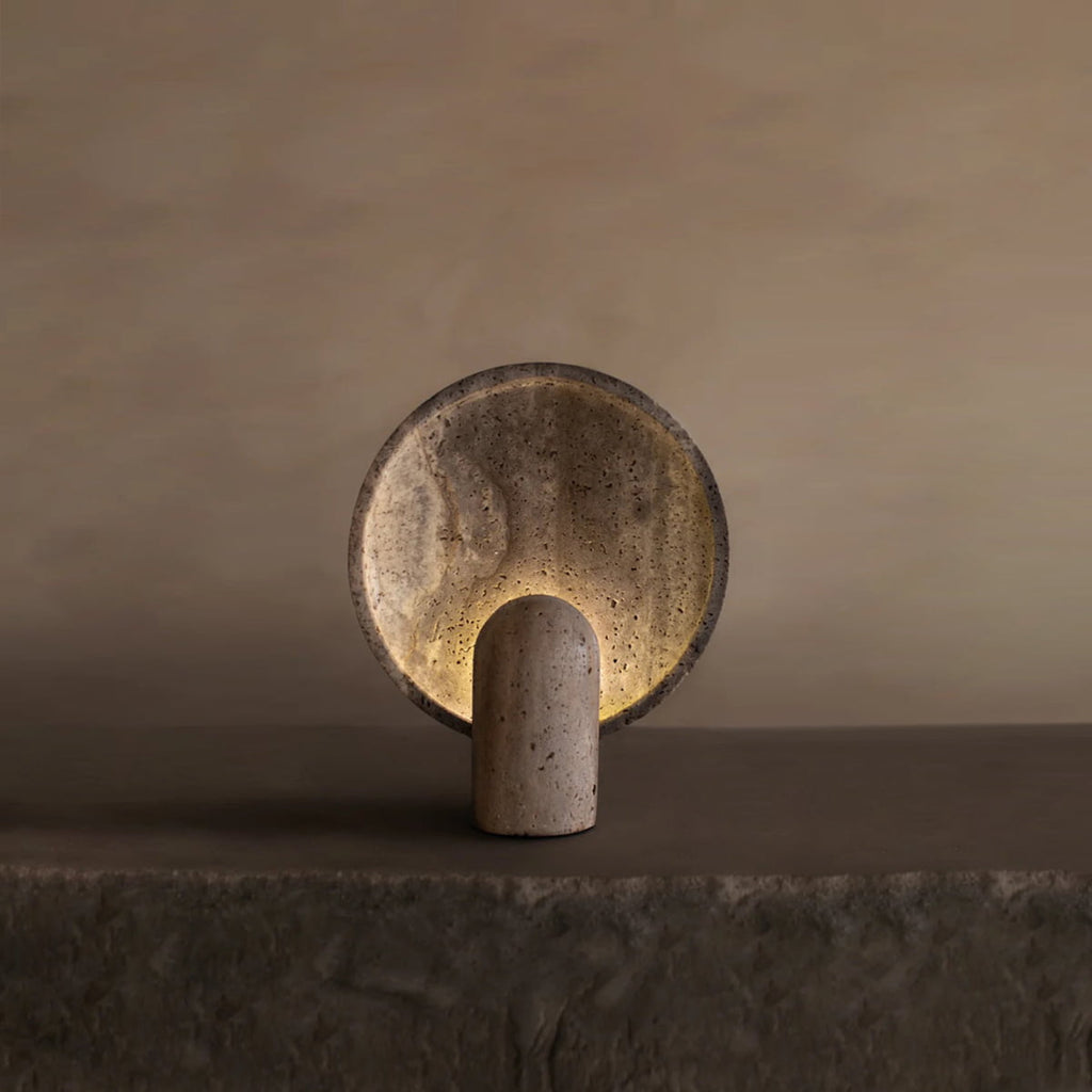 The Stella Lamp In Travertine features a round, travertine-inspired disc and cylindrical base, casting a soft, warm glow that enhances the serene ambiance of any space with its minimalist design and neutral tones.
