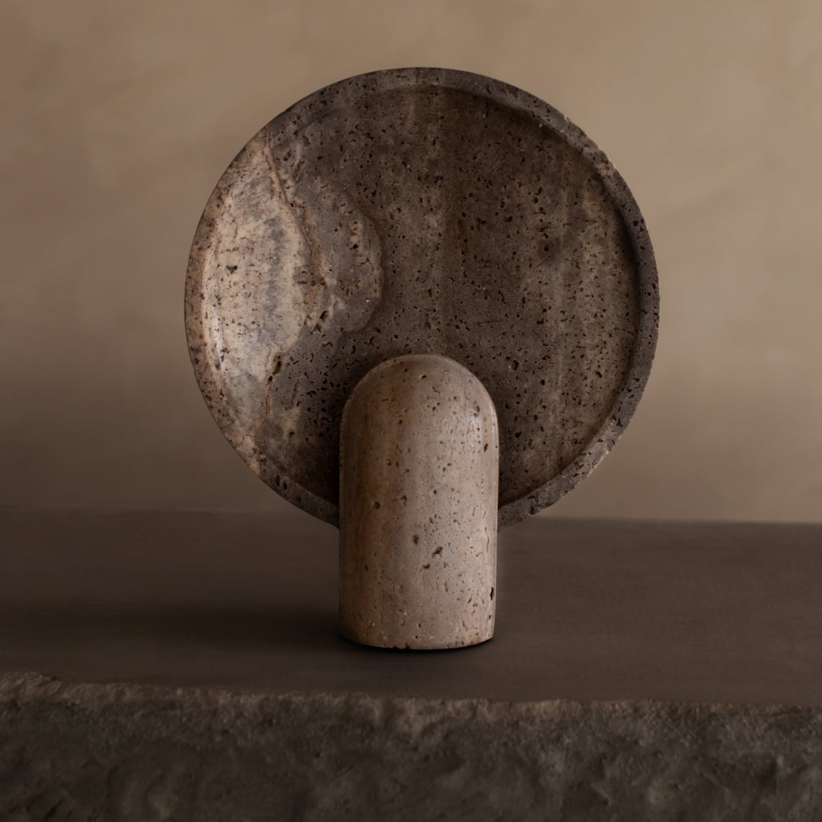 The Stella Lamp In Travertine features a smooth, dome-shaped travertine stone on a neutral-toned base, shown against a textured round plate and beige backdrop to create a calming, earthy ambiance.