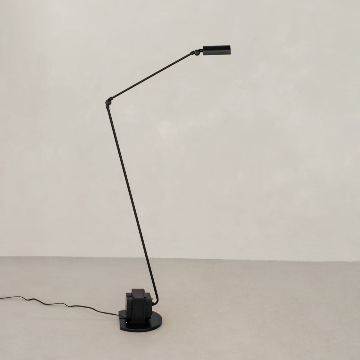 The black, modern Stem Lamp features a thin angled arm, cylindrical shade, and round base. Against a pale wall and floor, it offers adjustable height for versatile lighting, with the power cord lying neatly along the surface.