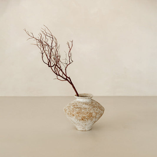 The Taru is a textured ceramic vase with a wide base and narrow neck, inspired by artisan pottery. Set against a light beige background, it elegantly holds a single bare brown branch—perfect for sophisticated home decor.