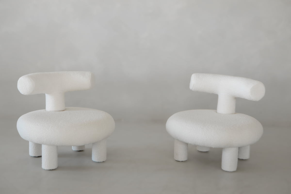 Two modern Teddy Chair Rental pieces with rounded seats, short cylindrical legs, and T-shaped backrests sit against a light gray background. Plush and minimalist, these sculptural chairs make a bold statement in any space.