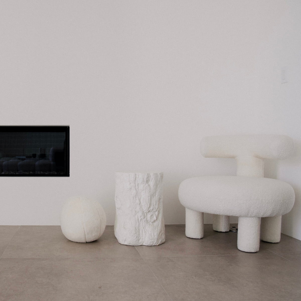 Minimalist room with a luxury Teddy Chair Rental, a textured white stool, and a round white cushion on gray tiled flooring beside a black-framed fireplace set in plain white walls.