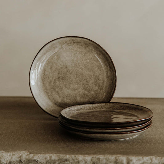 Four Tundra Glazed Dish Large ceramic plates with a rustic brown glaze are stacked on a textured stone surface, with one plate standing upright behind them against a neutral light beige background.