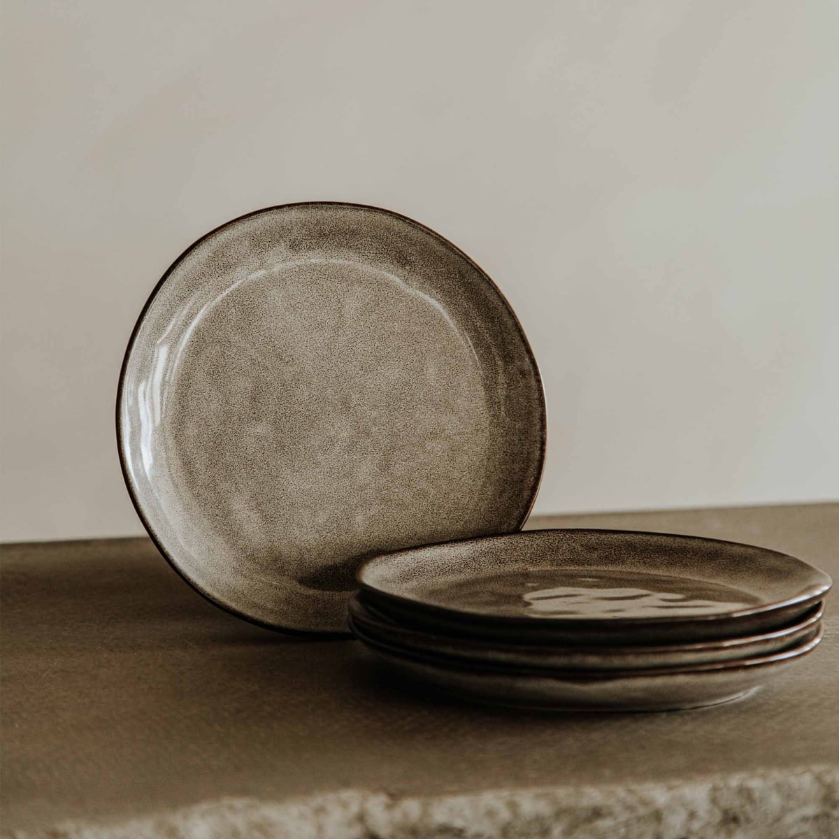 Four Tundra Glazed Dish Large plates with a rustic matte finish are stacked on a textured stone surface, with one plate standing upright behind the stack against a neutral background.