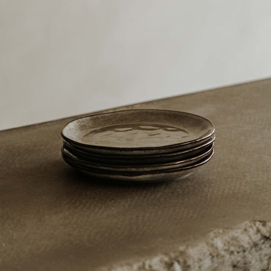 A stack of four Tundra Glazed Dish Small plates rests on a textured brown surface, set against a softly blurred neutral background.