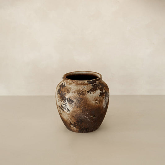 The Yogi vase, a rustic ceramic piece with a mottled brown and beige finish, sits on a neutral surface against a softly textured, pale background—showcasing handcrafted artisan charm.