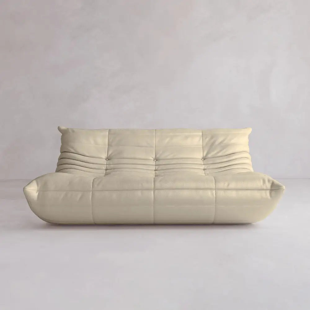 The Rolo Lounge Three Seater is a modern, minimalist beige sofa with soft cushions and subtle stitching, inspired by mid-century design, shown against a light gray textured wall in an empty room.