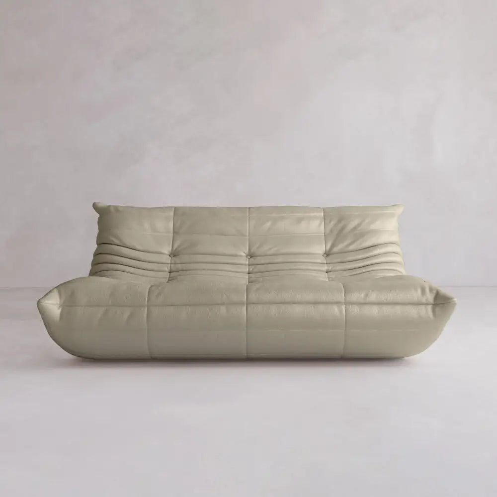 The Rolo Lounge Three Seater, a modern, low-profile beige sofa with tufted curved cushions and minimalist mid-century design, sits against a light textured wall in an empty room.