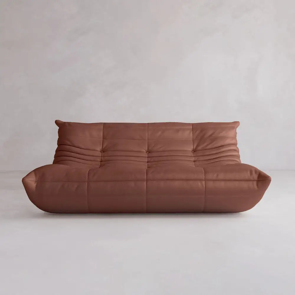 The Rolo Lounge Three Seater is a modern brown leather sofa with a low profile, deep seat, and soft quilted upholstery. It features subtle mid-century design elements and sits against a light-colored wall and floor.