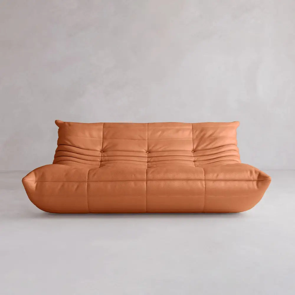 The Rolo Lounge Three Seater is a modern, low-profile tan leather sofa with a tufted, softly wrinkled surface in a mid-century design, shown against a plain light gray wall and floor.