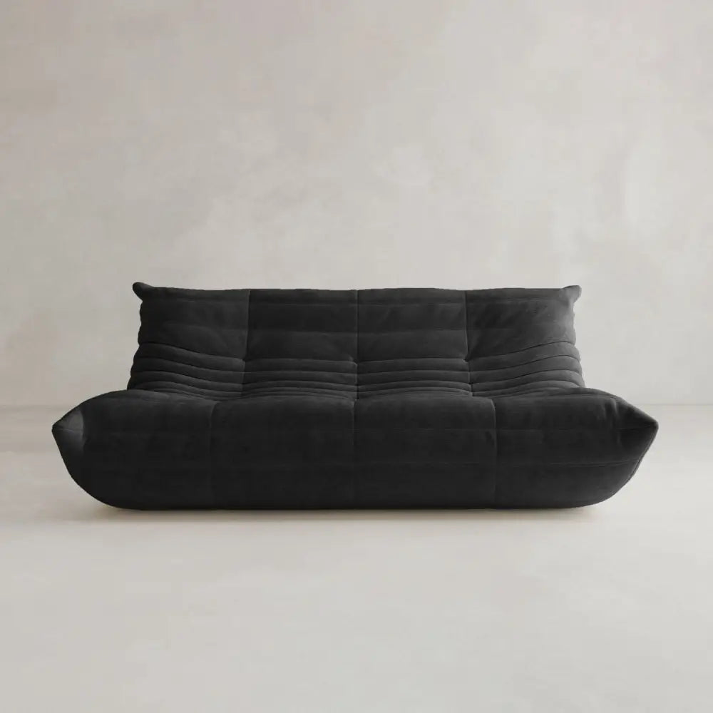 The Rolo Lounge Three Seater, a modern low-profile black sofa with tufted cushions inspired by mid-century design, is set against a light gray minimalist background.