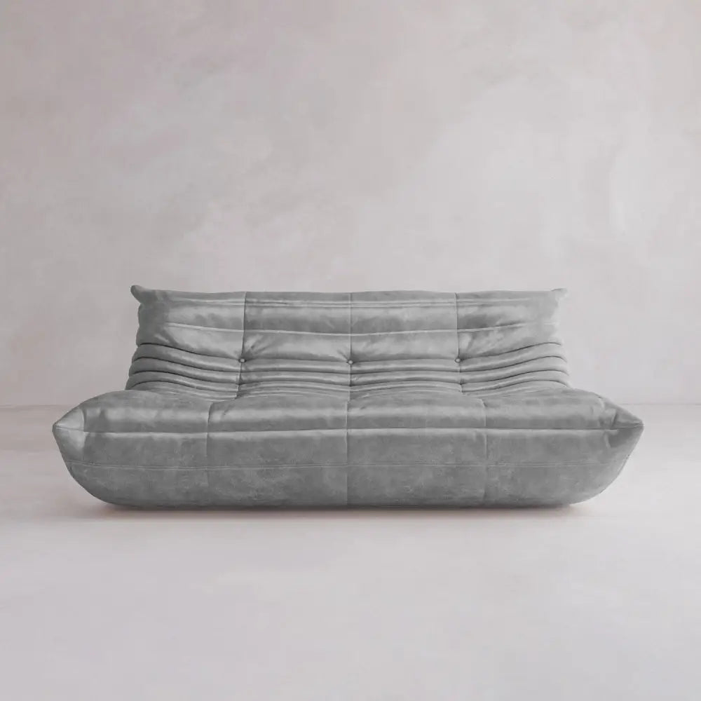 The Rolo Lounge Three Seater is a modern, low-profile gray leather sofa featuring a tufted, slightly wrinkled mid-century design, showcased against a light gray minimalist backdrop.