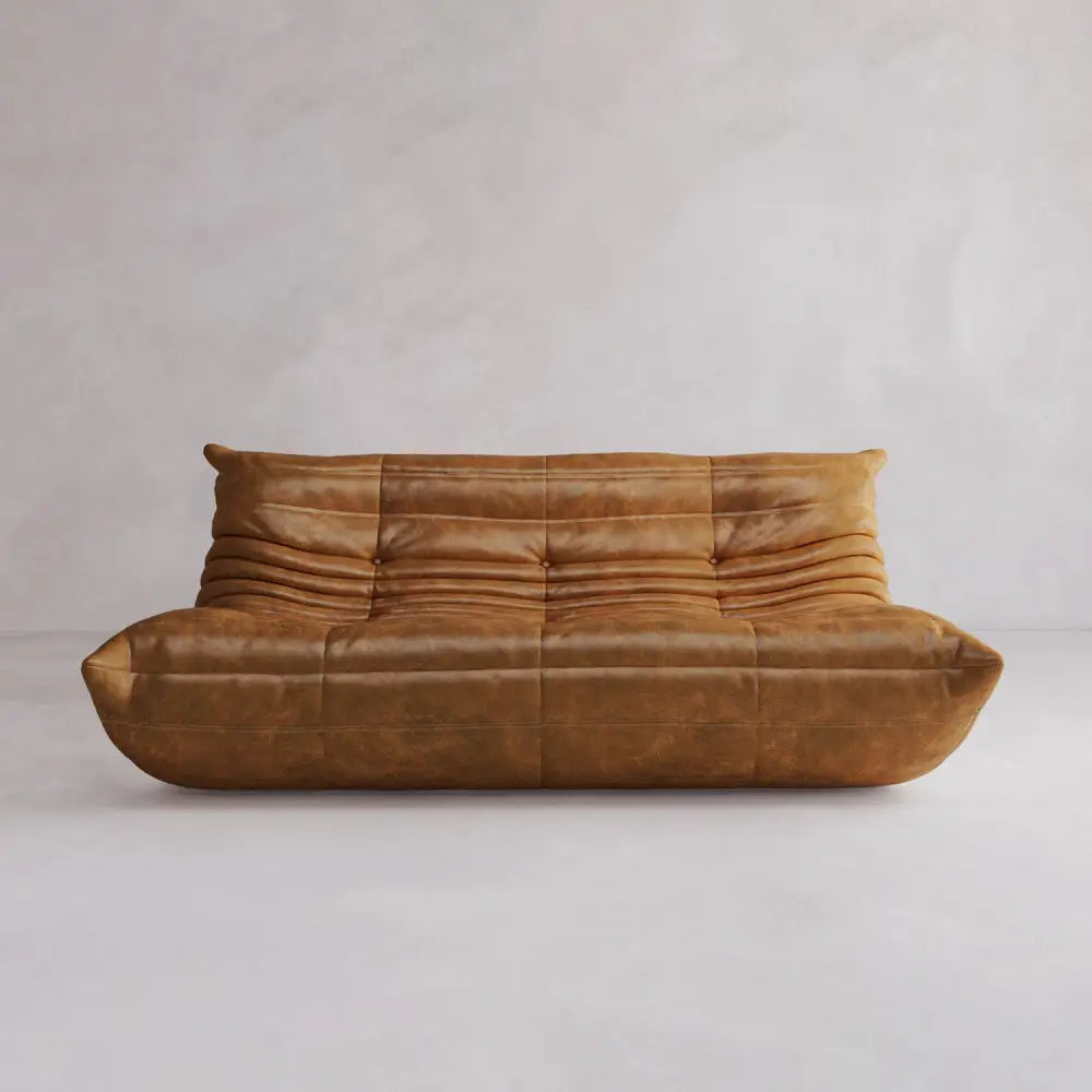 The Rolo Lounge Three Seater is a brown leather, tufted modular sofa with soft, rounded edges, shown against a plain light-colored wall and floor.