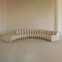 The Serpenti Sofa 15 Piece With Ottoman Rental is a large, curved beige leather modular system with tufted cushions and addable armchair elements for flexible seating, set against a plain light wall in a minimalist room.